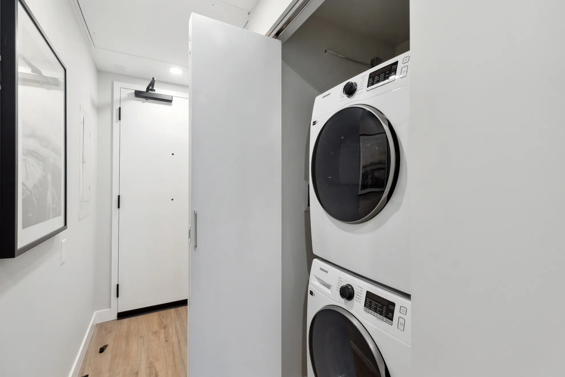 Bright apartment entryway featuring a stacked Samsung washer and dryer with white cabinetry and wood flooring.
